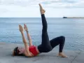 Mujer en posición de puente con una pierna elevada, practicando una técnica de carrera para mejorar la fuerza y el equilibrio junto al mar.