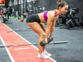 Mujer concentrada realizando un **double db deadlift** en un gimnasio, con pesas en cada mano y el cuerpo inclinado.