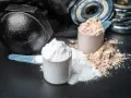 Dos cucharadas de proteína en polvo, una blanca y otra marrón, junto a pesas. ¡Cuidado! la proteína en polvo caduca.