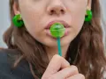 Labios rosados con un chupachups verde. Los pendientes son grandes y verdes. El maltitol es malo, pero este dulce parece tentador.