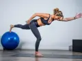 Mujer realizando ejercicios de fuerza sin material, en equilibrio sobre una pierna, con un balón azul y una caja negra al fondo.