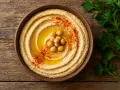 Un cremoso hummus en un cuenco de barro, decorado con garbanzos, aceite y pimentón. ¡Cuidado, el hummus engorda si no se consume con moderación!