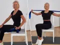 Mujer rubia realiza ejercicios en silla con banda elástica para tonificar brazos y piernas.