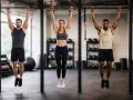 Tres personas, dos hombres y una mujer, se cuelgan de la barra en un gimnasio, entrenando su fuerza.