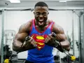 Hombre con camiseta de Superman haciendo un curl bayesian en el gimnasio, con expresión de esfuerzo.