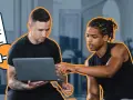 Entrenador y cliente revisan un programa de entrenamiento en una laptop. Un icono de lista de tareas pendientes con "PLAN" destaca.