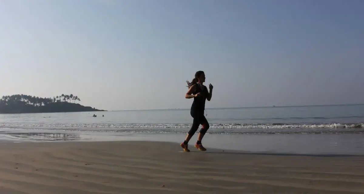 Mujer corriendo en la playa al amanecer, con el mar y una isla con palmeras al fondo.