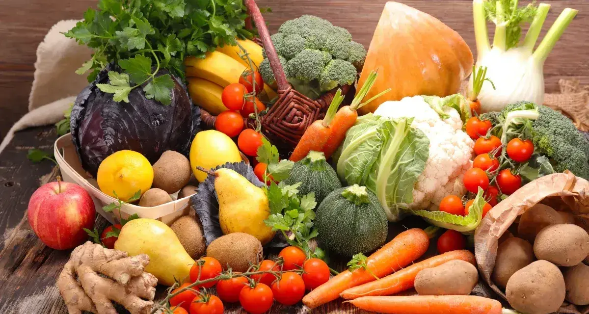 Variedad de tipos de verdura y frutas frescas sobre mesa de madera: brócoli, coliflor, zanahorias, tomates, repollo morado, peras, manzanas, plátanos, limones, jengibre y patatas.