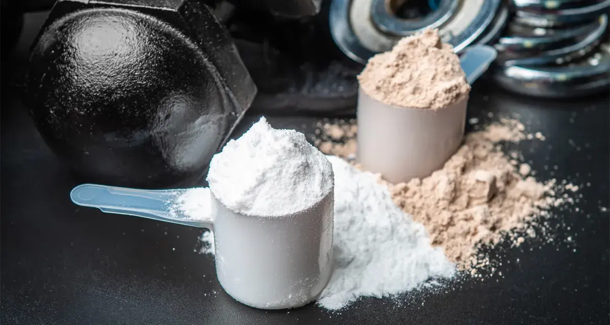 Dos cucharadas de proteína en polvo, una blanca y otra marrón, junto a pesas. ¡Cuidado! la proteína en polvo caduca.
