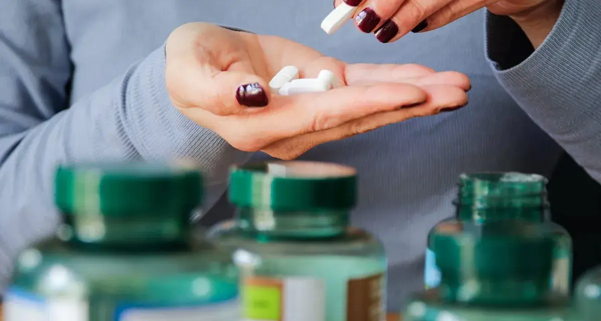 Manos con uñas oscuras sostienen pastillas blancas. Se ven frascos de suplementos. Descubre los beneficios del magnesio en el hombre.