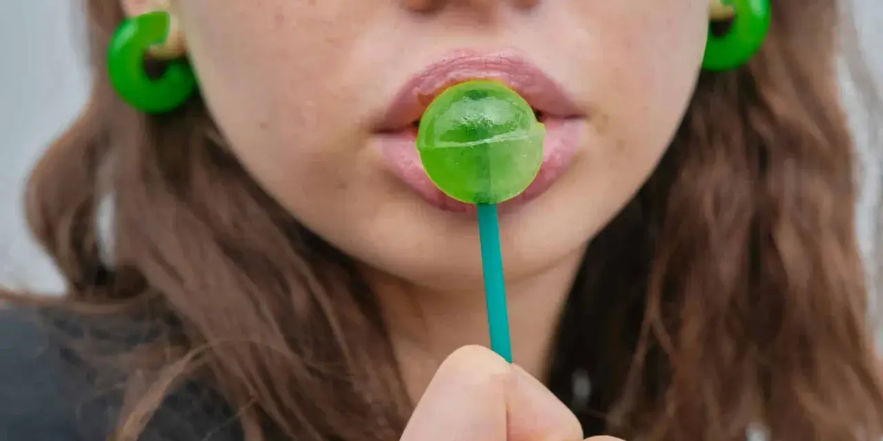 Labios rosados con un chupachups verde. Los pendientes son grandes y verdes. El maltitol es malo, pero este dulce parece tentador.