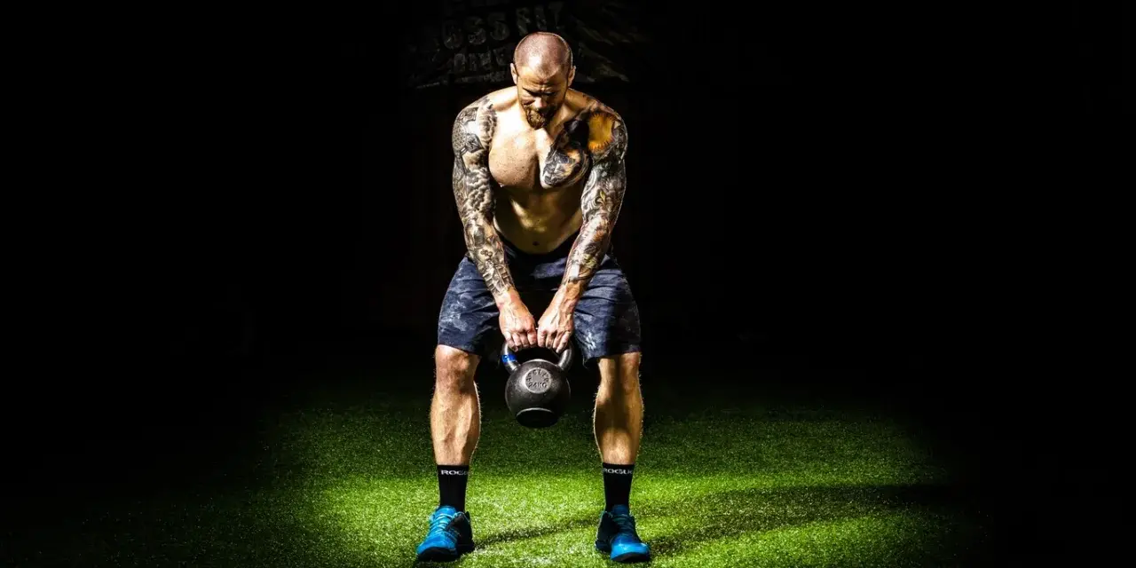 Hombre tatuado realiza press militar con kettlebell, enfocado en su entrenamiento.