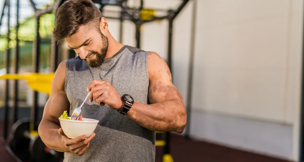 Hombre con barba en gimnasio, comiendo ensalada. Demuestra que se puede ganar músculo en déficit calórico con la dieta adecuada.