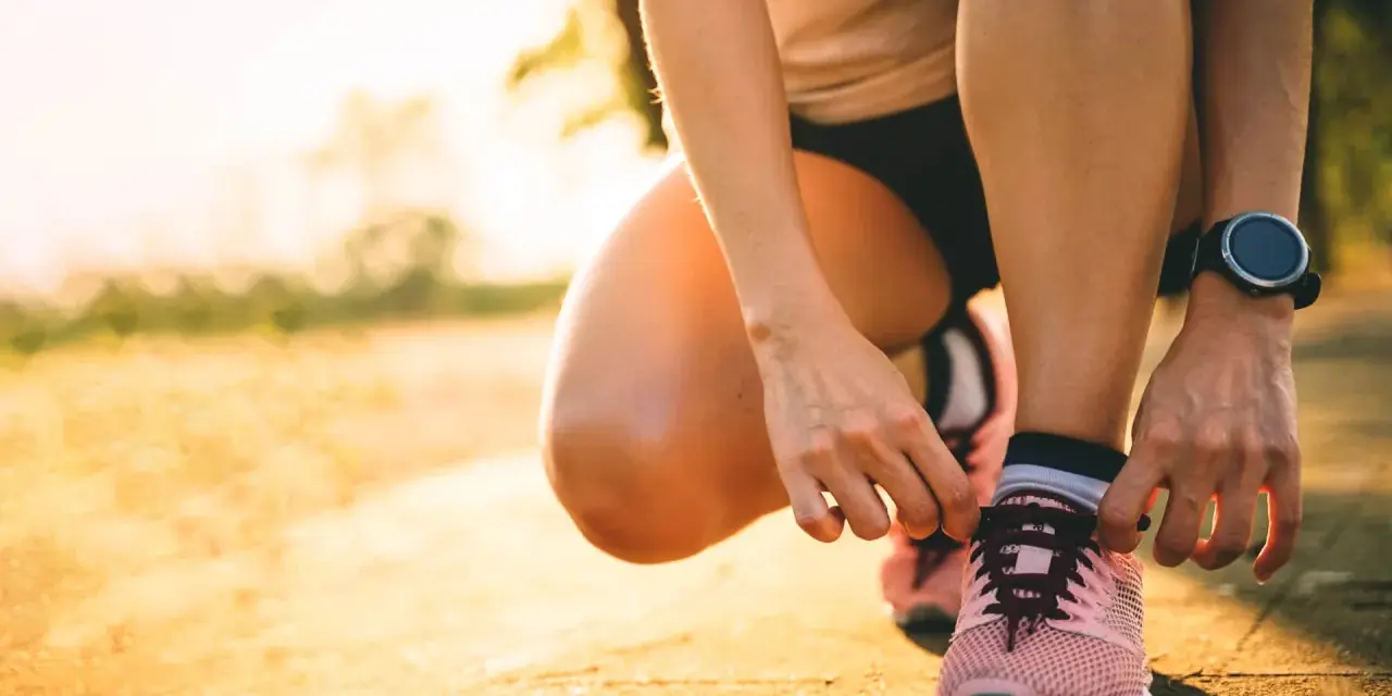 Persona se agacha para atarse los cordones de sus zapatillas rosas, lista para salir a correr al amanecer.