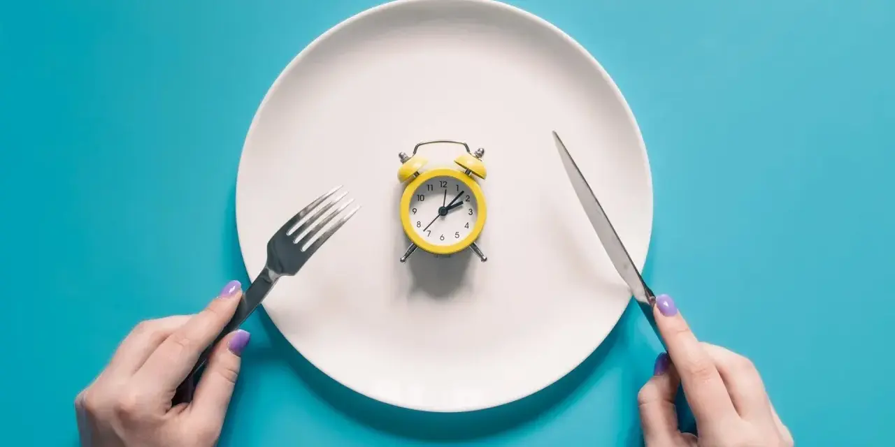 Plato blanco con despertador amarillo en el centro. Manos sostienen tenedor y cuchillo. El café rompe el ayuno.