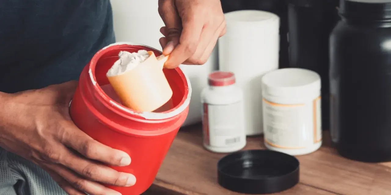 Persona prepara su suplemento. Se muestra un recipiente rojo con polvo blanco y una cuchara. Varios envases de suplementos en el fondo. Calculando la dosis creatina por peso.