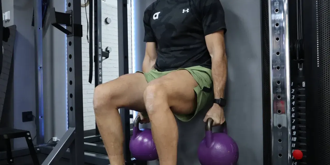 Hombre realizando tabla de ejercicios isométricos, sentado contra la pared con pesas rusas moradas.