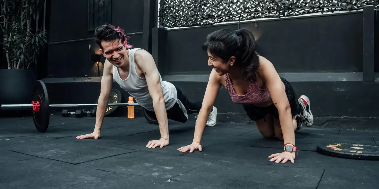 Pareja sonríe haciendo flexiones, un ejemplo de cuales son los ejercicios de fuerza.