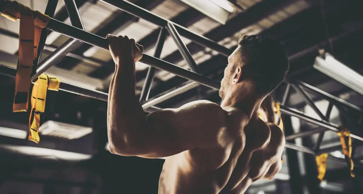 Hombre musculoso realizando pull ups en un gimnasio. Sus brazos y espalda están tensos mientras se eleva.
