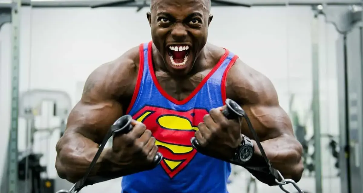 Hombre con camiseta de Superman haciendo un curl bayesian en el gimnasio, con expresión de esfuerzo.