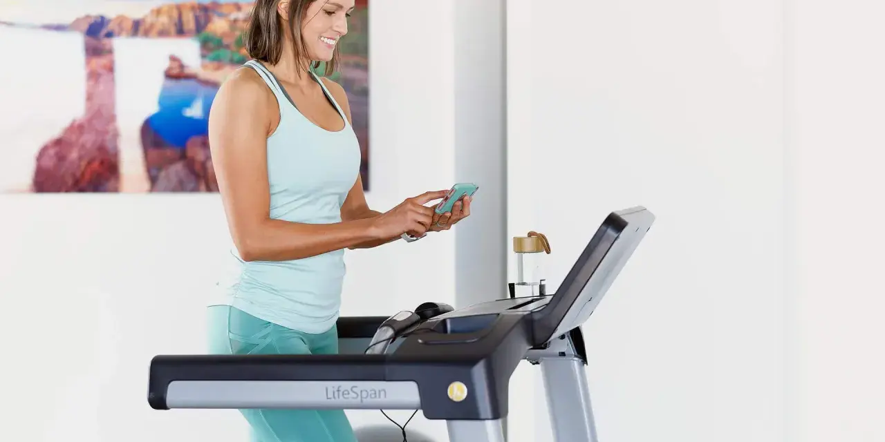 Mujer sonriente en cinta de correr, revisando su teléfono. Quizás buscando cuántas calorías debo comer al día para mantener su energía.