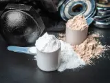 Dos cucharadas de proteína en polvo, una blanca y otra marrón, junto a pesas. ¡Cuidado! la proteína en polvo caduca.