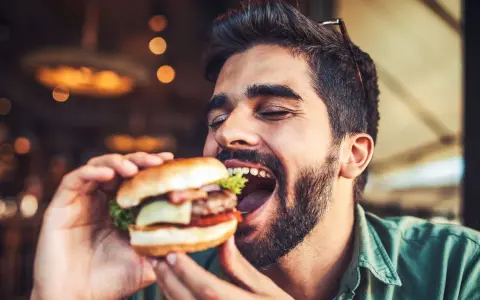 Hombre disfrutando un jugoso **cheat meal** de hamburguesa, con los ojos cerrados por el placer.