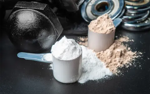 Dos cucharadas de proteína en polvo, una blanca y otra marrón, junto a pesas. ¡Cuidado! la proteína en polvo caduca.