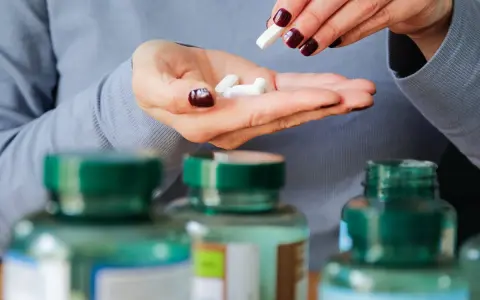 Manos con uñas oscuras sostienen pastillas blancas. Se ven frascos de suplementos. Descubre los beneficios del magnesio en el hombre.
