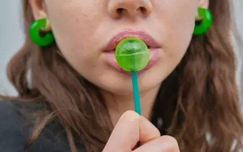 Labios rosados con un chupachups verde. Los pendientes son grandes y verdes. El maltitol es malo, pero este dulce parece tentador.