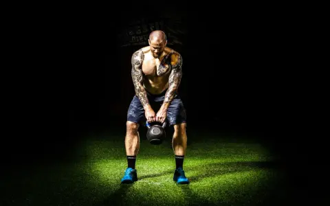 Hombre tatuado realiza press militar con kettlebell, enfocado en su entrenamiento.