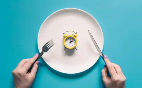 Plato blanco con despertador amarillo en el centro. Manos sostienen tenedor y cuchillo. El café rompe el ayuno.