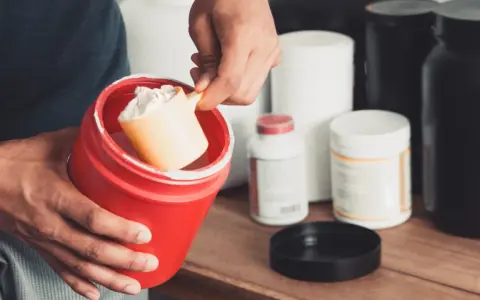 Persona prepara su suplemento. Se muestra un recipiente rojo con polvo blanco y una cuchara. Varios envases de suplementos en el fondo. Calculando la dosis creatina por peso.