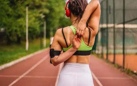 Mujer con cuerpo tonificado estirando el brazo sobre la cabeza en una pista de atletismo.