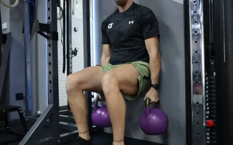 Hombre realizando tabla de ejercicios isométricos, sentado contra la pared con pesas rusas moradas.