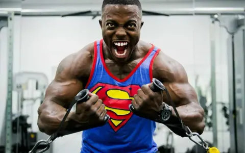 Hombre con camiseta de Superman haciendo un curl bayesian en el gimnasio, con expresión de esfuerzo.