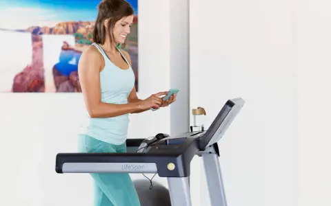 Mujer sonriente en cinta de correr, revisando su teléfono. Quizás buscando cuántas calorías debo comer al día para mantener su energía.