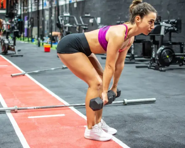 Mujer concentrada realizando un **double db deadlift** en un gimnasio, con pesas en cada mano y el cuerpo inclinado.