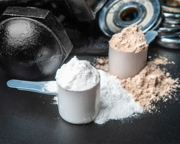 Dos cucharadas de proteína en polvo, una blanca y otra marrón, junto a pesas. ¡Cuidado! la proteína en polvo caduca.