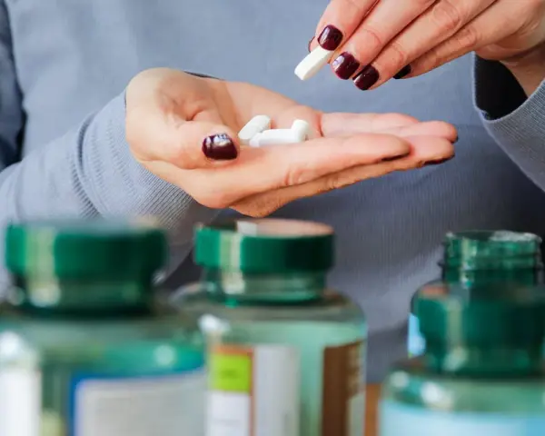 Manos con uñas oscuras sostienen pastillas blancas. Se ven frascos de suplementos. Descubre los beneficios del magnesio en el hombre.