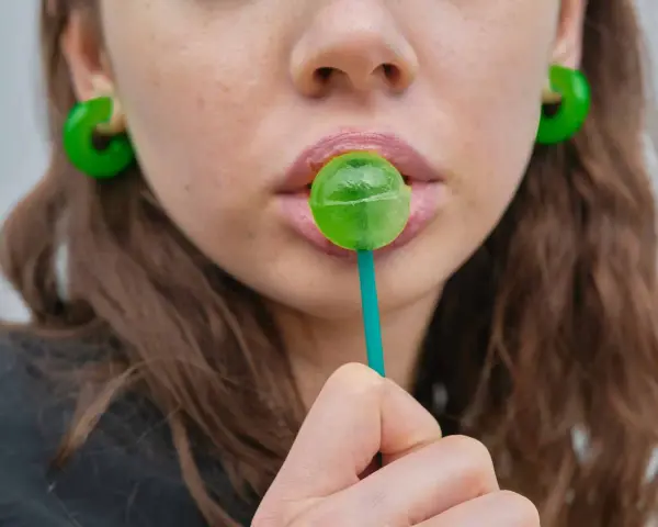 Labios rosados con un chupachups verde. Los pendientes son grandes y verdes. El maltitol es malo, pero este dulce parece tentador.