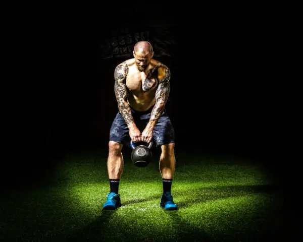 Hombre tatuado realiza press militar con kettlebell, enfocado en su entrenamiento.