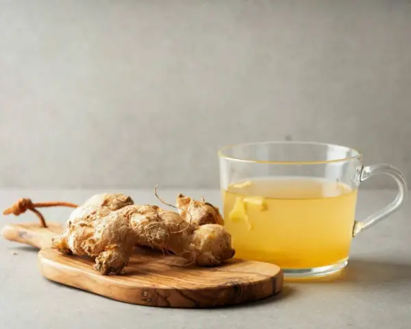 Raíz de jengibre en tabla de madera junto a una taza de té de jengibre, destacando sus propiedades beneficiosas.