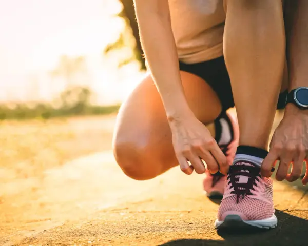 Persona se agacha para atarse los cordones de sus zapatillas rosas, lista para salir a correr al amanecer.