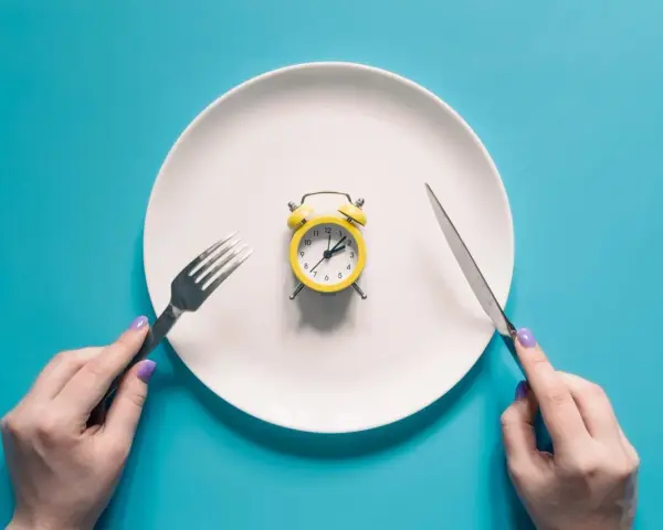 Plato blanco con despertador amarillo en el centro. Manos sostienen tenedor y cuchillo. El café rompe el ayuno.