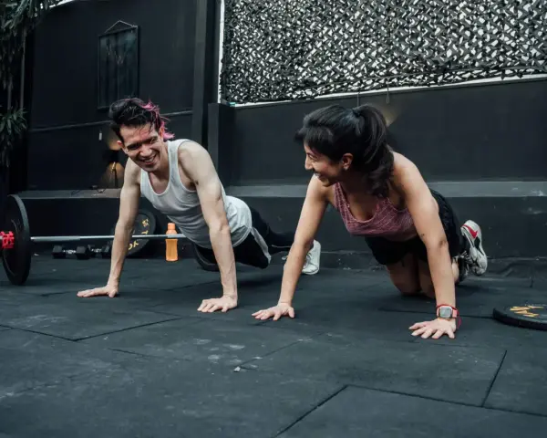 Pareja sonríe haciendo flexiones, un ejemplo de cuales son los ejercicios de fuerza.