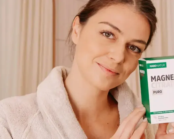 Mujer sonriente con bata, sosteniendo una caja de Magnesio Citrato Puro. Una dosis de magnesio para evitar calambres y mejorar la energía.