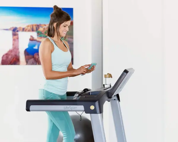 Mujer sonriente en cinta de correr, revisando su teléfono. Quizás buscando cuántas calorías debo comer al día para mantener su energía.