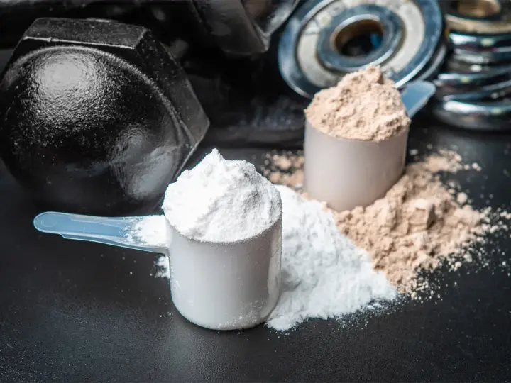 Dos cucharadas de proteína en polvo, una blanca y otra marrón, junto a pesas. ¡Cuidado! la proteína en polvo caduca.