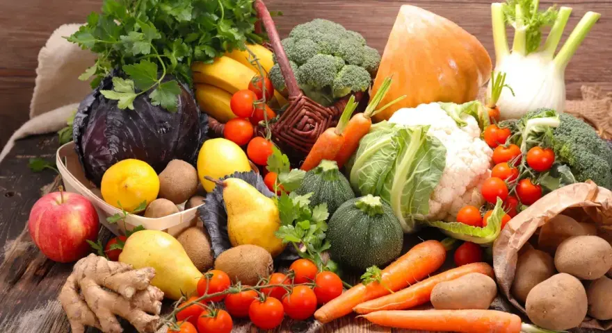 Variedad de tipos de verdura y frutas frescas sobre mesa de madera: brócoli, coliflor, zanahorias, tomates, repollo morado, peras, manzanas, plátanos, limones, jengibre y patatas.